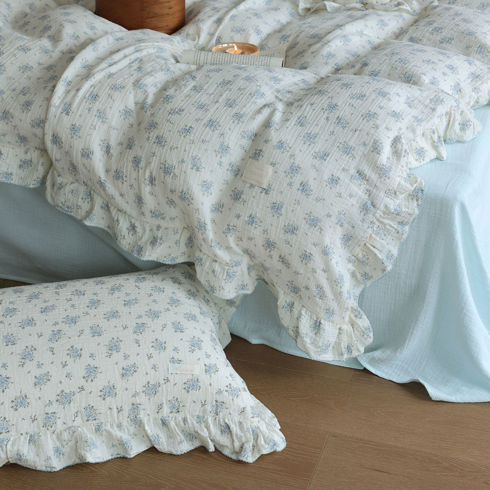 A lower-angle view of a queen-sized bed featuring a blue floral cotton muslin duvet cover set with matching ruffled pillow. The soft, wrinkled texture of the muslin fabric is visible, draped over a solid blue flat sheet.