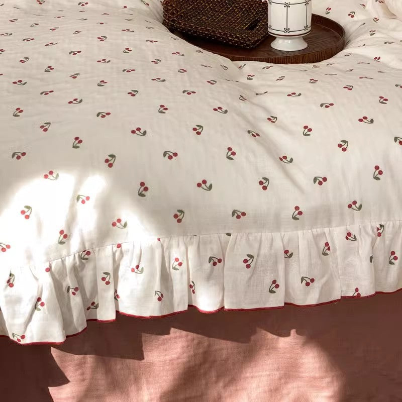Close-up texture shot of a breathable cotton muslin bedding set featuring a repeating tiny cherry print pattern and a detailed view of the decorative ruffle trim.