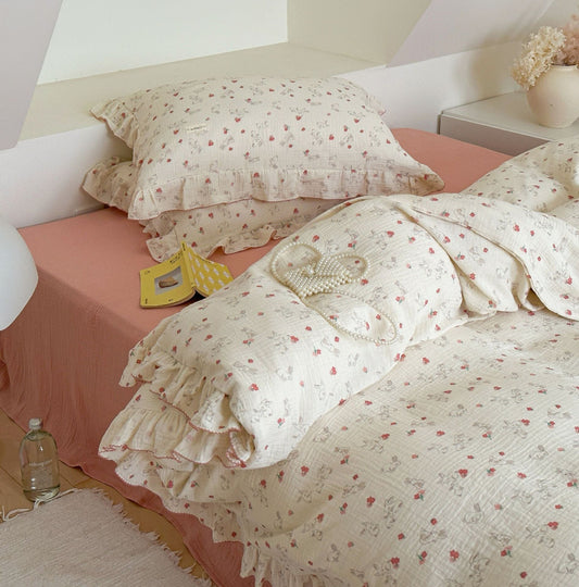 An over-the-bed shot of a cotton duvet cover set with a whimsical rabbit and floral print. The image captures the cozy, relaxed feel of the bedding, with its ruffled edges and a plush toy. A subtle perfume bottle and a pearl necklace on the bed suggest a luxurious, modern style.