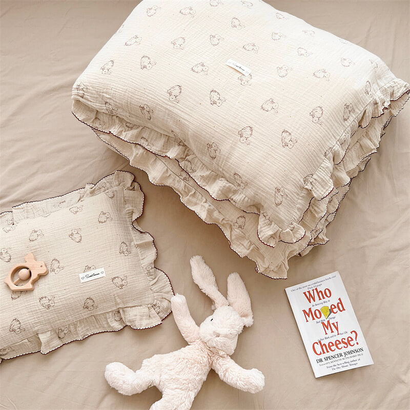 An overhead shot of a cotton ruffle duvet set with a bunny print, showcasing a folded duvet and a pillow. The bedding has a soft, wrinkled texture and ruffled edges with contrasting stitching. A plush bunny toy and a book are on the bed, hinting at a cozy and playful style.