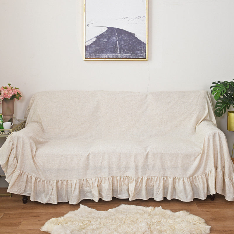 A beige ruffled sofa slipcover on a three-seater sofa, with a large painting on the wall and a potted plant nearby, showing the full length of the cover.