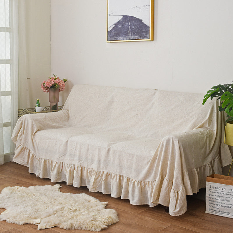 The beige ruffled sofa cover on a couch in a living room, with a soft rug on the floor and a large piece of wall art, providing a clear view of the entire slipcover.