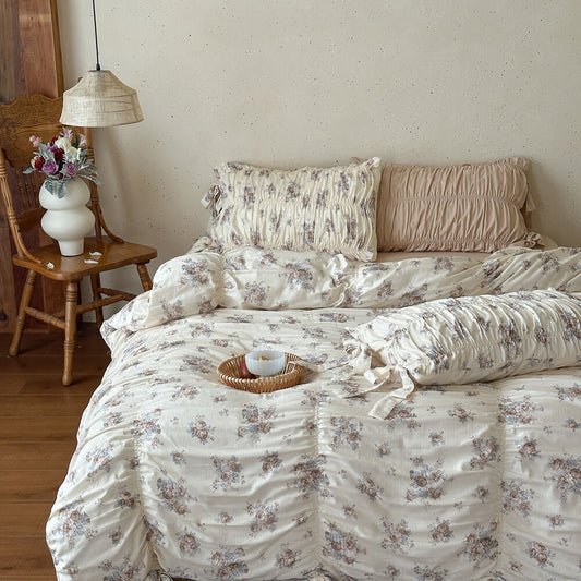 Cream cotton muslin bedding set with faded rose floral print, ruched pillow shams, ribbon ties, and beige sheet styling.