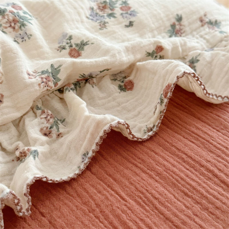 A detailed close-up shot of a floral duvet cover with a ruffled edge, placed on a pink bedsheet. The image focuses on the intricate floral pattern in shades of blue, green, and pink, and the soft texture of the cotton gauze fabric. The ruffled edge is highlighted with contrasting dark stitching.