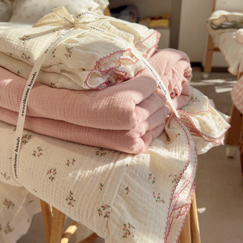 A close-up of a stack of a pink floral print cotton muslin bedding set tied with a decorative ribbon. The image highlights the ruffled edges with a light pink border and shows layers of both the patterned and solid-colored bedding, emphasizing the quality and soft texture.