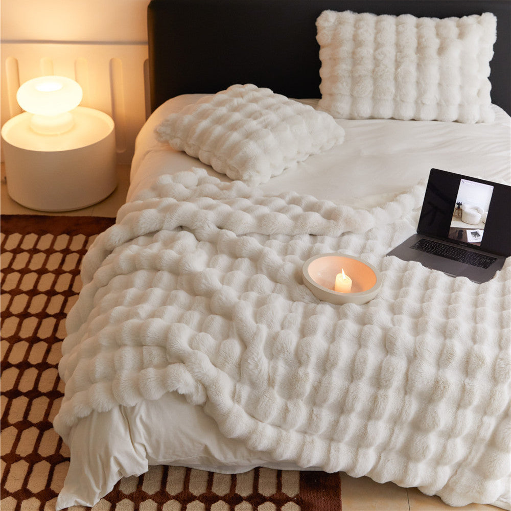A cozy, white faux rabbit fur throw blanket with a bubbly texture, draped over a bed. A matching pillow and a laptop are on the bed.