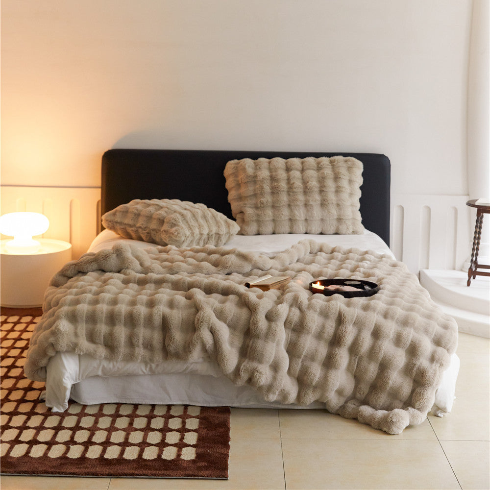 A warm, tan-colored faux rabbit fur throw blanket with a puffy grid texture, covering a bed with a matching pillow.