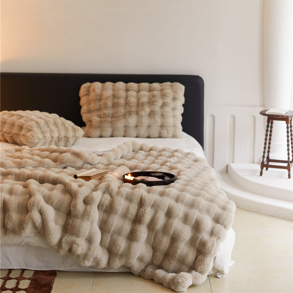 An overall view of a bed styled with a soft, tan-colored faux rabbit fur throw blanket and matching pillows.