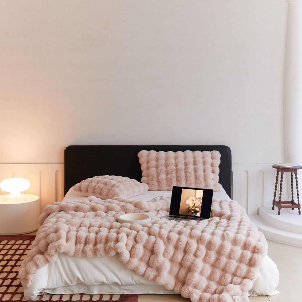 A cozy, blush pink faux rabbit fur throw blanket with a bubble pattern, styled on a bed with a matching pillow.