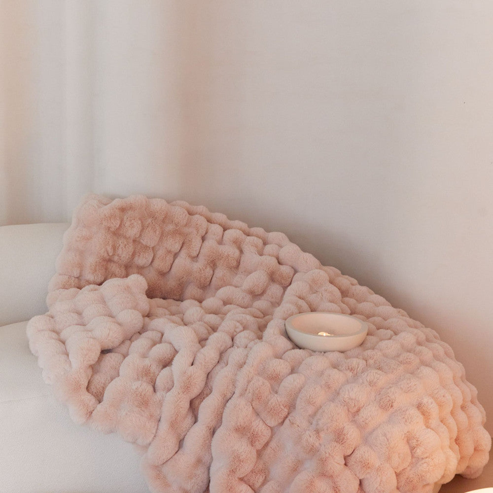A close-up of a soft, blush pink faux rabbit fur blanket with a bubbly texture, draped over a sofa.