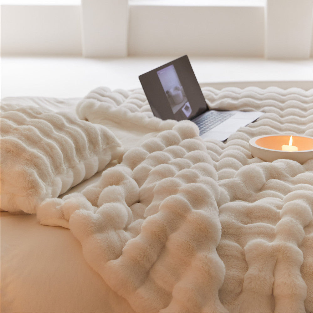 A close-up shot of a white, fluffy faux rabbit fur blanket with a bubbly texture on a bed, with a pillow and laptop in the background.