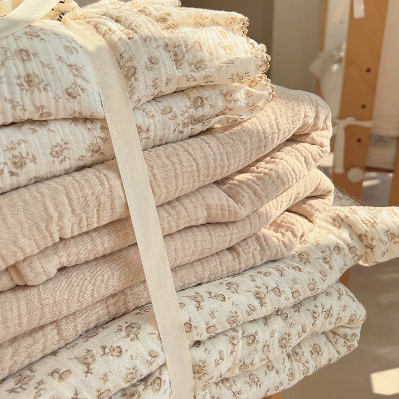 A close-up of a neatly folded vintage cotton muslin bedding set, showcasing a stack of the brown camellia floral duvet cover and a solid khaki-colored layer. The soft, textured fabric and the ruffled edges with brown stitching are clearly visible.