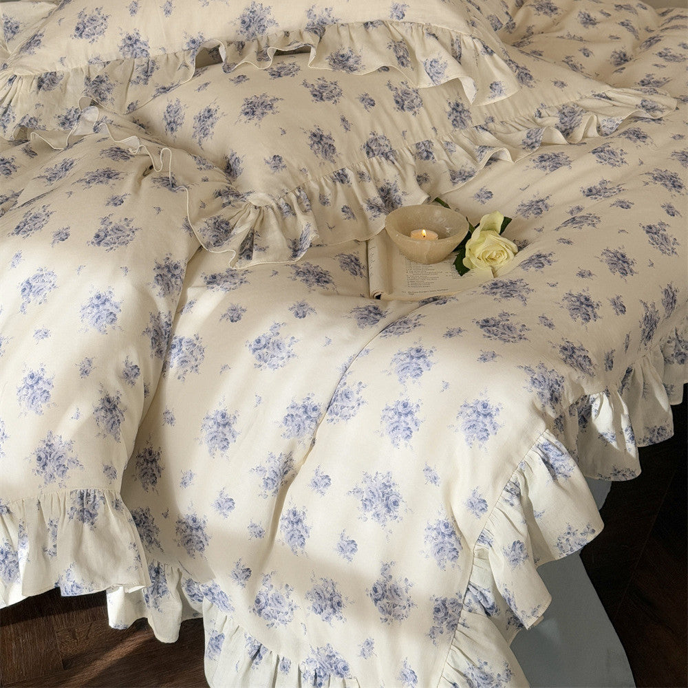Close-up: Blue rose floral ruffled bedding, styled with an open book, small candle bowl, and a white rose.