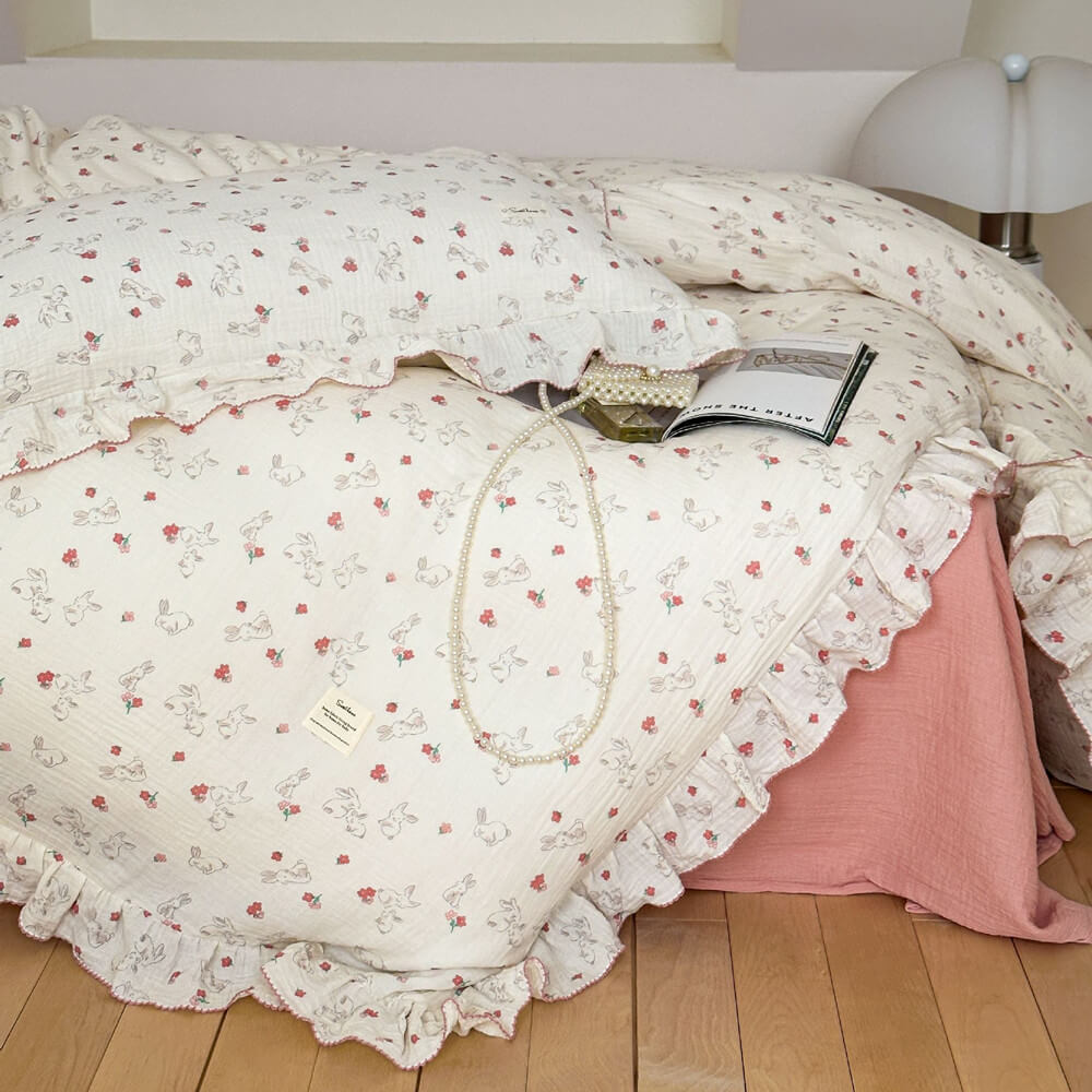 A full-bed shot of a rabbit and floral print cotton muslin duvet cover set. The image shows the full bedding with ruffled edges, a pink flat sheet, and a matching pillow. A strand of pearls and an open book add to the romantic and serene aesthetic of the bedroom.