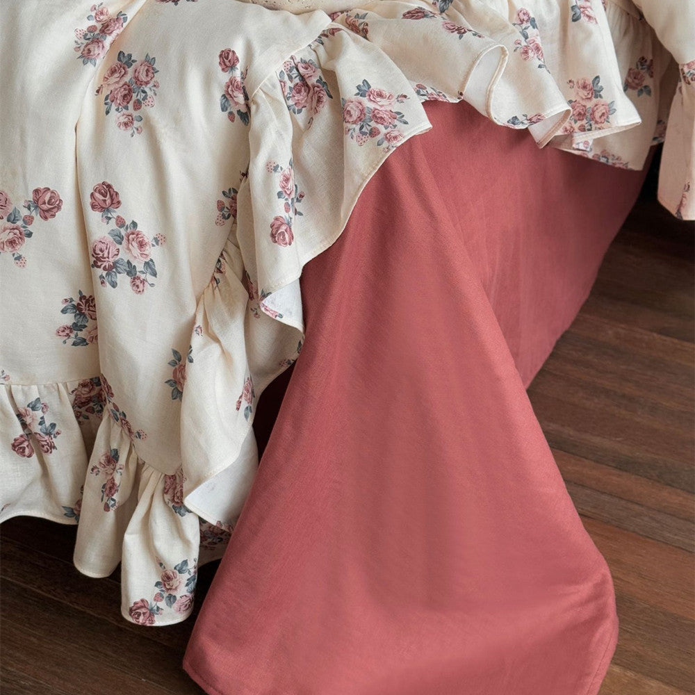 Corner close-up showing the contrast between the floral ruffled cotton duvet and the solid rust flat sheet on a hardwood floor.