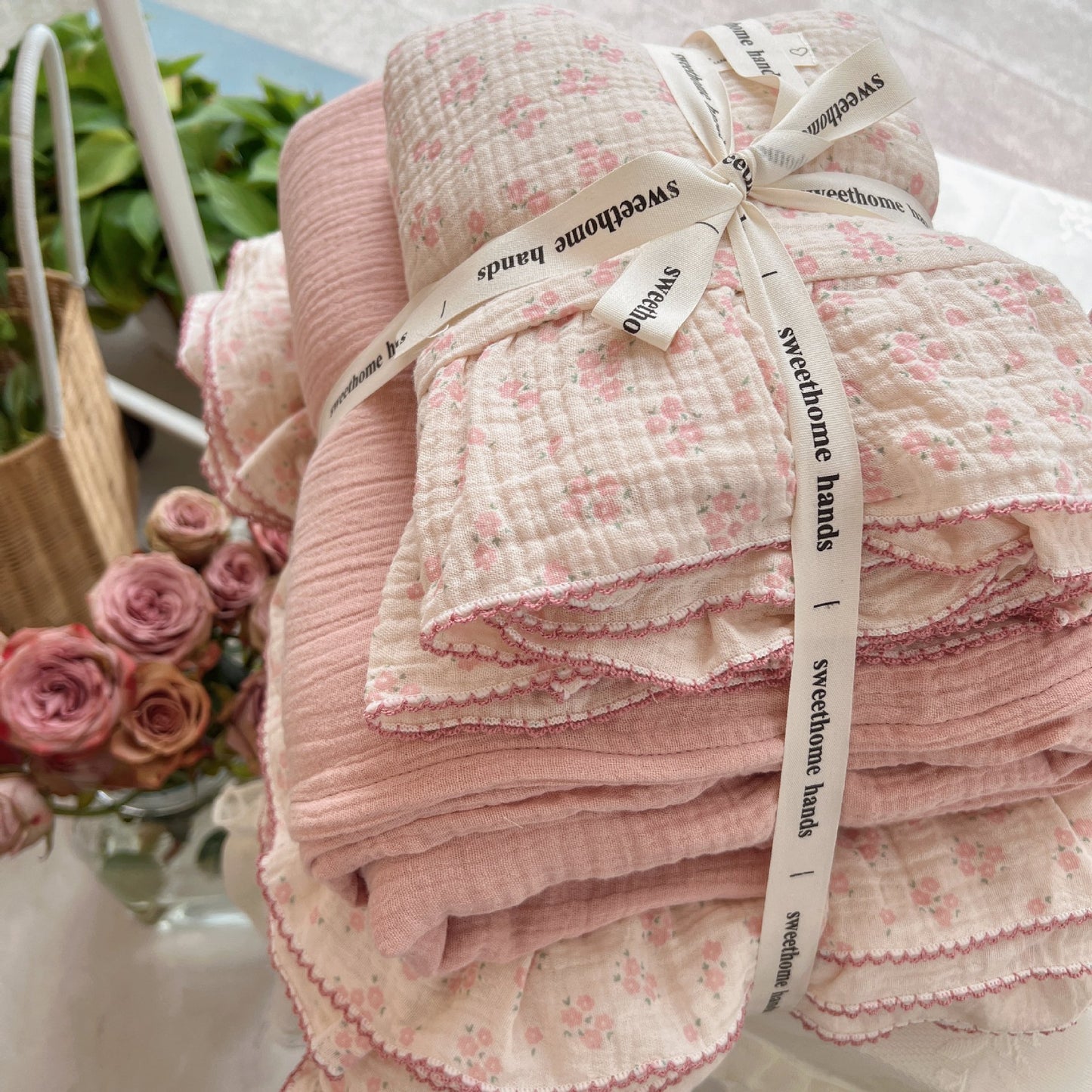 A neatly folded pink floral cotton muslin duvet cover set tied with a decorative ribbon, showcasing the ruffled edges and the soft, textured fabric. The bedding stack includes both the light pink floral pattern and a solid pink layer, with a bouquet of pink roses visible in the background.