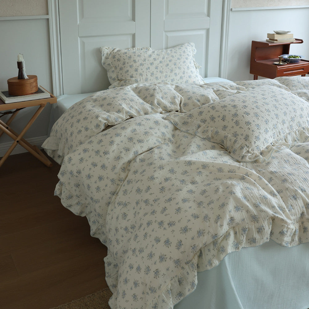 A full view of a cozy bed dressed with a blue floral cotton muslin bedding set. The duvet cover and pillow shams are a light off-white with scattered blue flowers and ruffle edges.