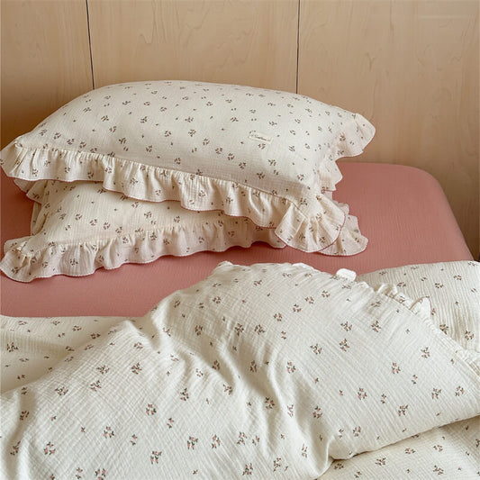 A close-up of a delicate floral cotton gauze duvet cover set on a bed with a pink flat sheet. Two ruffled pillowcases are stacked, showing the soft fabric texture and subtle floral print. The image focuses on the quality and detailed design of the bedding.