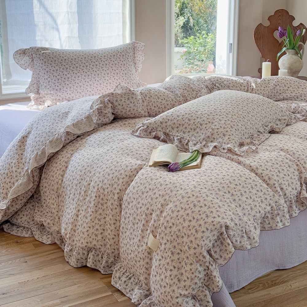 A bedroom setting with a purple floral duvet cover set on a bed, accompanied by a flat sheet and pillowcases. The bedding set has a naturally crinkled texture and is displayed in a room with a window and a book with a purple cover on the bed.