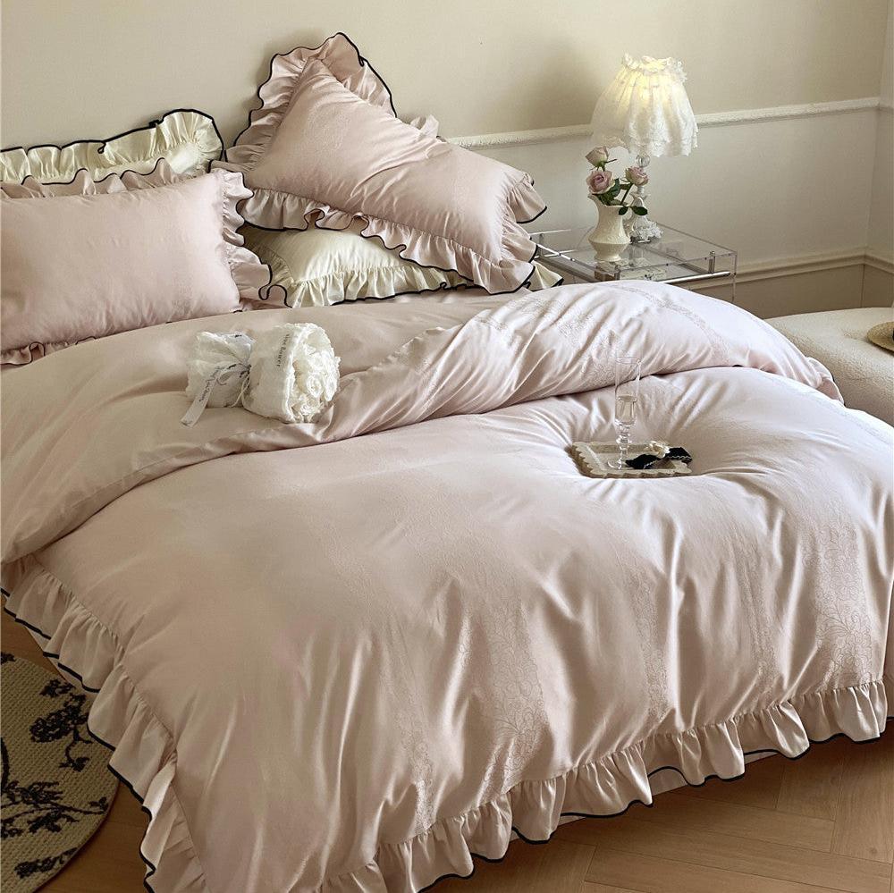 Full bed view of the silky ruffle bedspread set in light pink, showing a cozy arrangement with decorative glass and a white ruffled lamp.