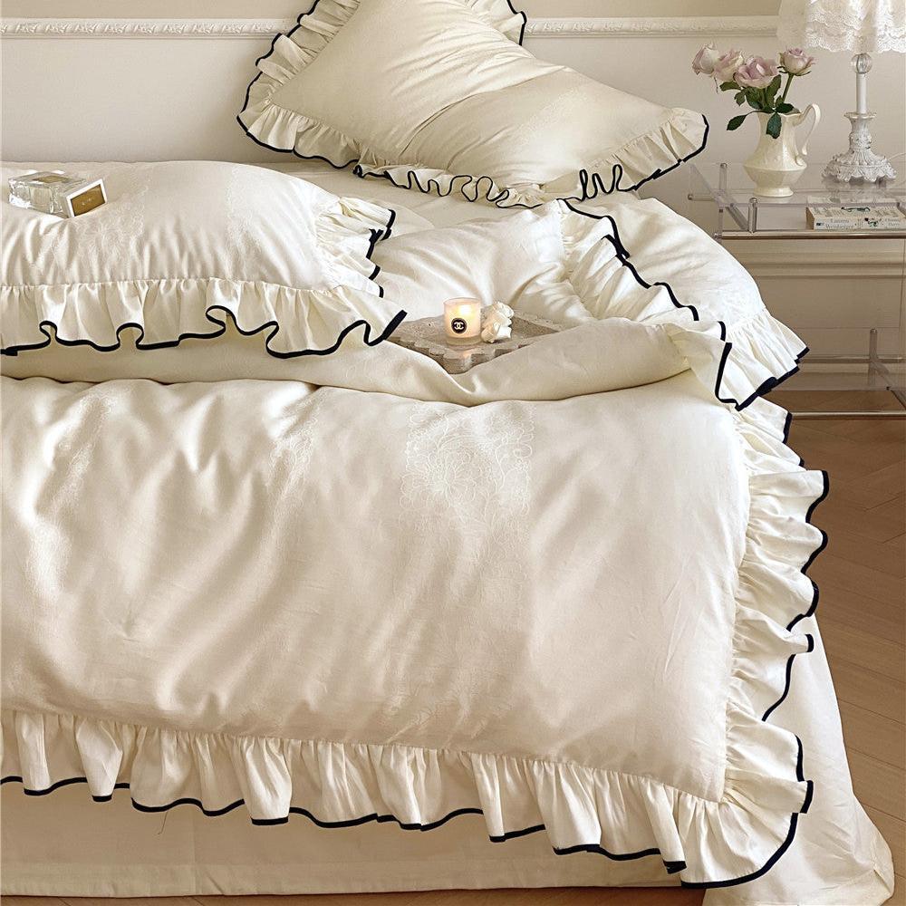 Full bed view of the cream white silky ruffle sham set with black trim, styled with a lace lamp and light pink roses on a bedside table.