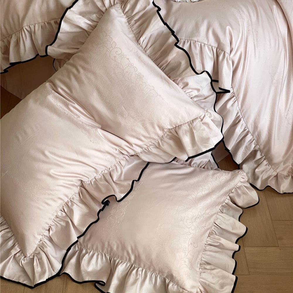Closeup detail of the smooth ruffle bedding set in light pink, showing two pillows on the floor with black trim and a subtle jacquard pattern.