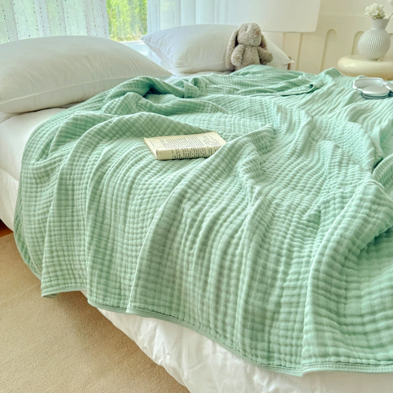 A soft sage green 6-layer cotton gauze blanket laid out as a throw on a bed in a sunny room.