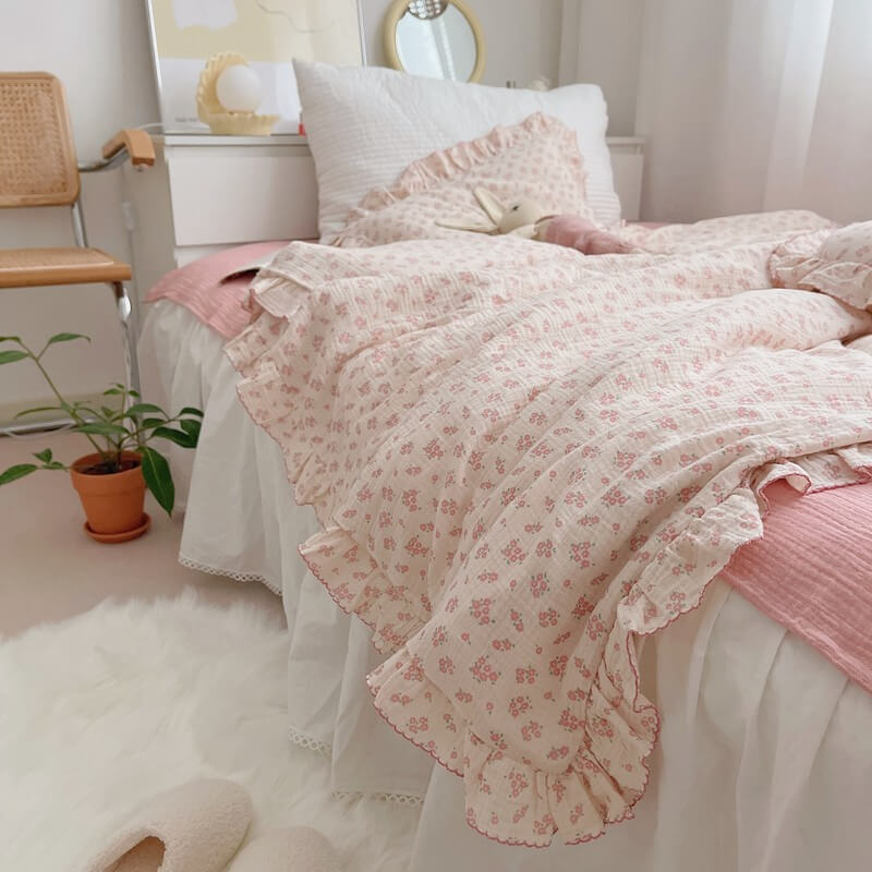 A side-angle view of a twin-size bed with a pink floral muslin duvet cover set. The image highlights the ruffles on the duvet and pillowcases, creating a layered and textured look. The white bed skirt with lace trim and the fluffy white rug add to the sweet, feminine charm of the girl's bedding.