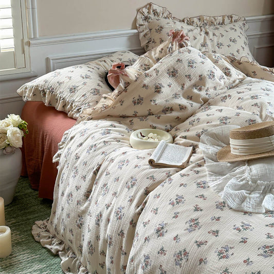 A floral duvet cover on a bed, surrounded by pillows, a book, a hat, and some candles on the floor.