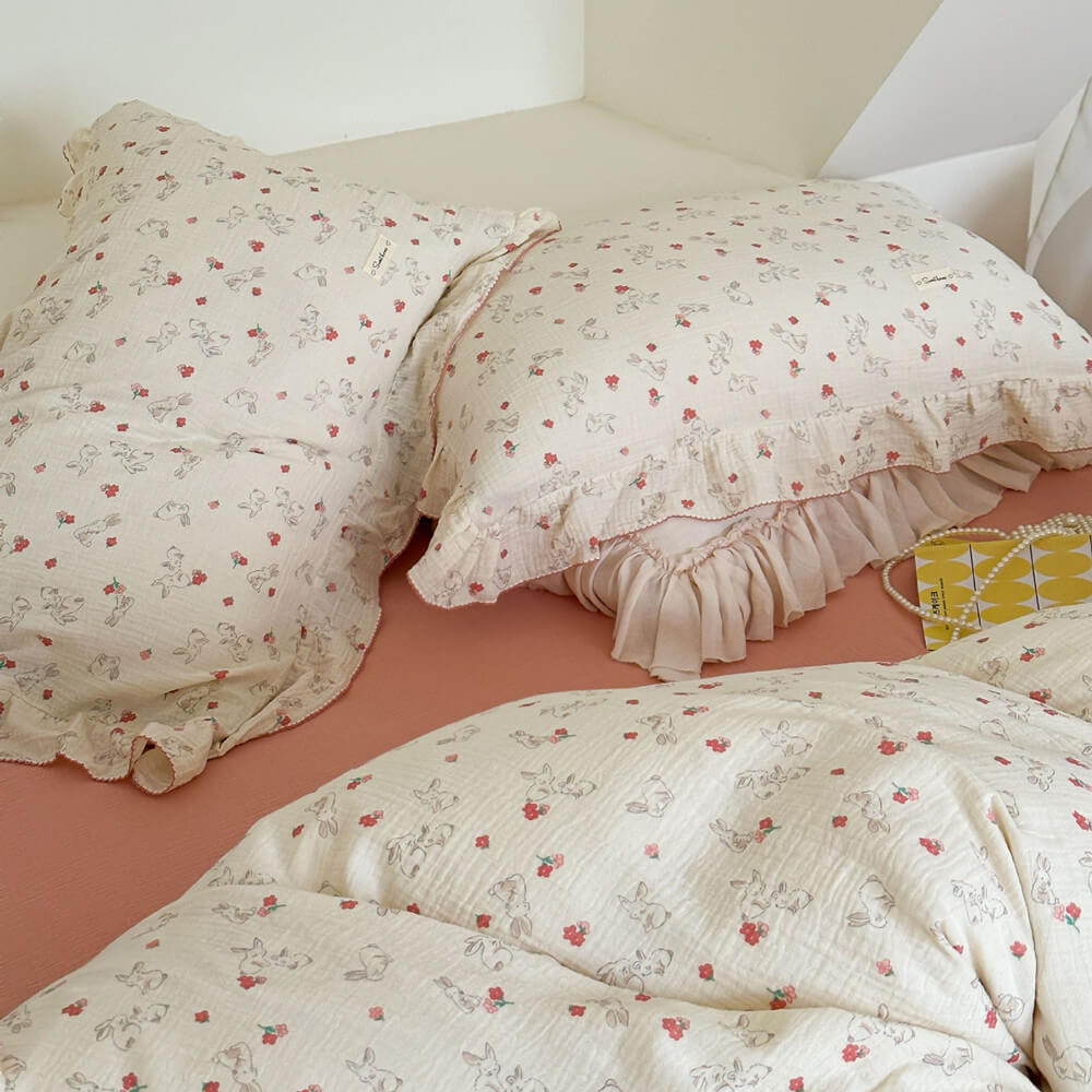 A side-angle view of a cozy bed featuring a cotton muslin duvet cover set with a rabbit and floral print. The bedding has elegant ruffled edges and is paired with a pink flat sheet and a lace-trimmed white bed skirt, creating a soft and feminine look for a girl's bedroom.