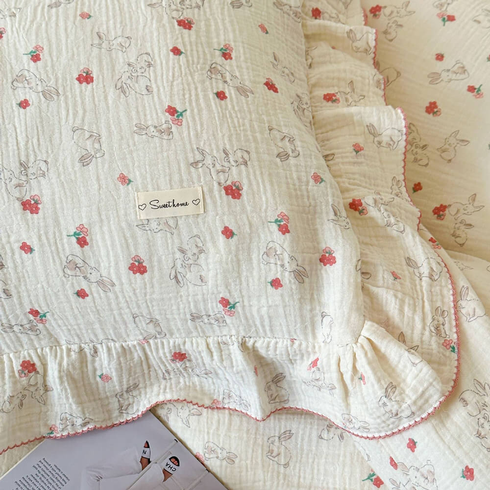 A close-up shot of a cotton muslin duvet cover set with a charming rabbit and floral print. The image focuses on a pillow, showcasing the soft fabric, delicate pink ruffles, and the unique, whimsical pattern of rabbits and tiny flowers.