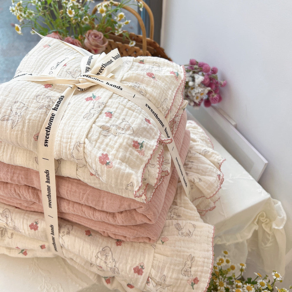 A close-up of a neatly folded cotton muslin duvet cover set tied with a decorative ribbon. The stack includes both the rabbit and floral print duvet and a solid pink layer. The image highlights the soft, crinkled texture of the fabric and the ruffled edges.