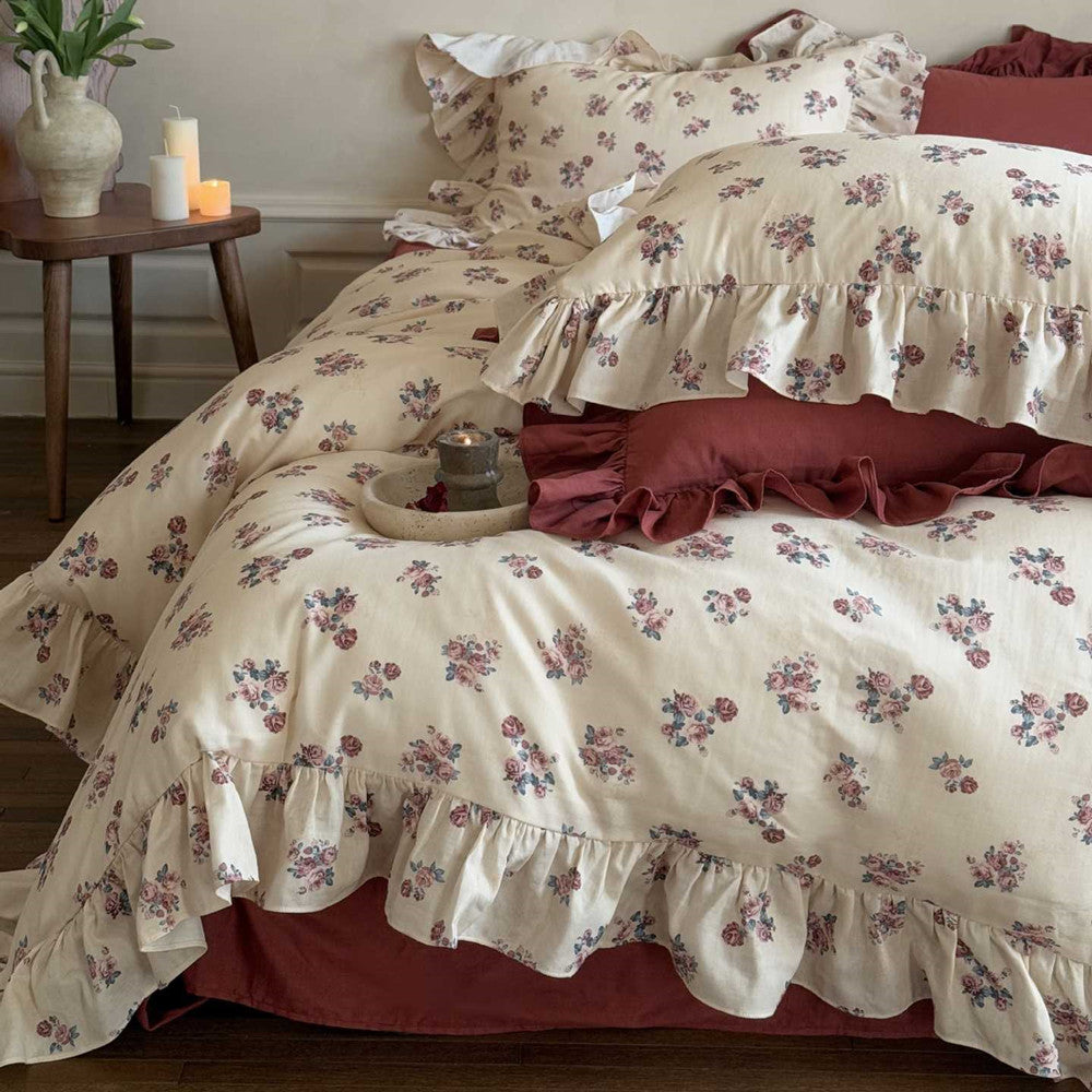Bed close-up showing the Vintage Rose Floral Bedding Set with a ruffled cotton design, accented by solid burgundy throw pillows and a bedside table.