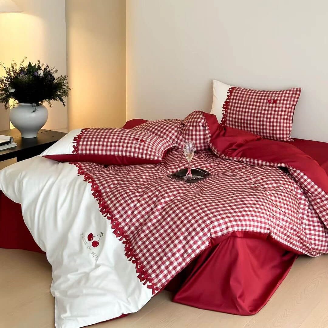 Cozy bedroom scene with red and white gingham cotton bedding set featuring cherry embroidery, white pillow, and bedside table with vase and lamp