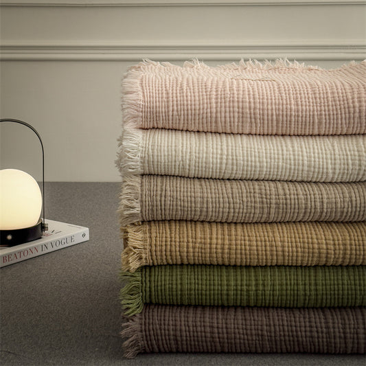Stack of six layered cotton muslin throw blankets in soft pastel and earth tones with fringed edges on gray surface by modern lamp