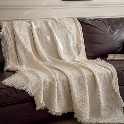 Cream-colored 6-layer cotton muslin throw blanket with fringed edges draped over a brown leather sofa