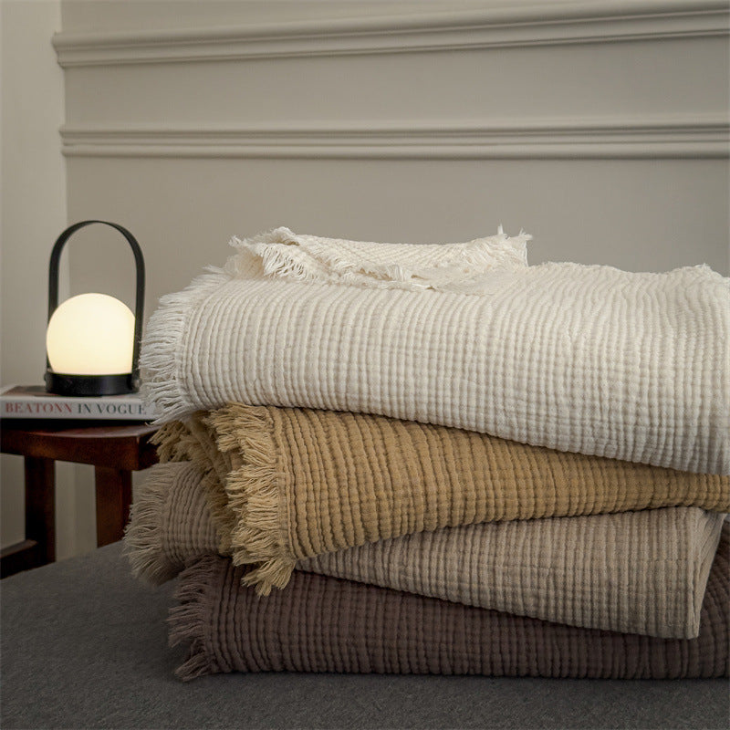 Stack of four soft 6-layer cotton muslin throw blankets in neutral tones with fringed edges on a grey surface next to a glowing lamp