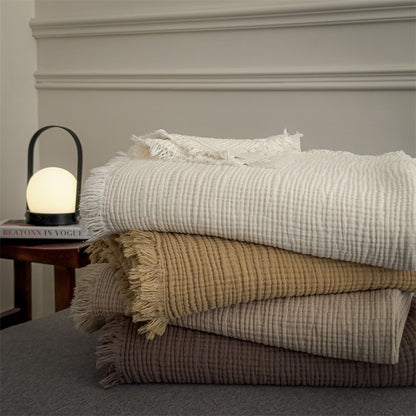 Stack of four soft 6-layer cotton muslin throw blankets in neutral tones with fringed edges on a grey surface next to a glowing lamp