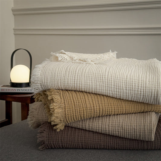 Stack of four soft 6-layer cotton muslin throw blankets in neutral tones with fringed edges on a grey surface next to a glowing lamp