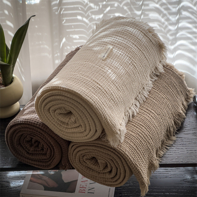 Three rolled 6-layer cotton muslin throw blankets in beige and brown on dark surface with potted plant and magazine