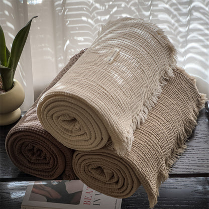 Three rolled 6-layer cotton muslin throw blankets in beige and brown on dark surface with potted plant and magazine