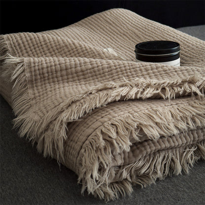 Beige 6-layer cotton muslin throw blanket with textured weave and fringed edges on dark fabric surface