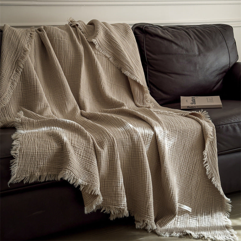 Beige 6-layer cotton muslin throw blanket with fringe draped over dark brown leather sofa with book
