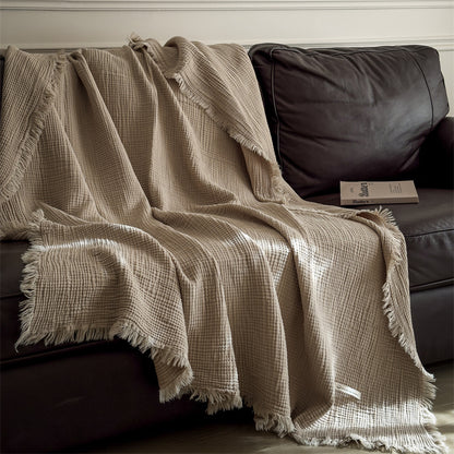 Beige 6-layer cotton muslin throw blanket with fringe draped over dark brown leather sofa with book