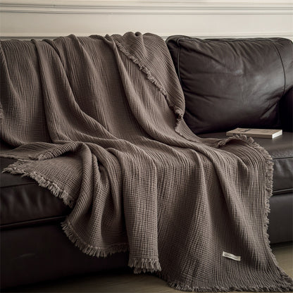 Dark brown 6-layer cotton muslin throw blanket draped over a black leather sofa with a book on the armrest