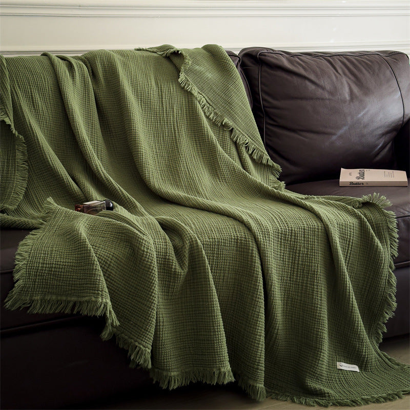 Green 6-layer cotton muslin throw blanket with fringed edges draped over dark leather sofa with book