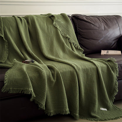 Green 6-layer cotton muslin throw blanket with fringed edges draped over dark leather sofa with book