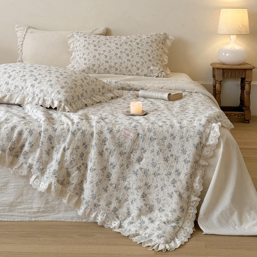 Blue rose comforter draped across a neutral bed with white lace edging and bedside lamp