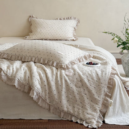 Shabby chic cream muslin comforter featuring a delicate blueberry botanical pattern and embroidered ruffle lace borders, displayed next to a rustic marble flower vase.
