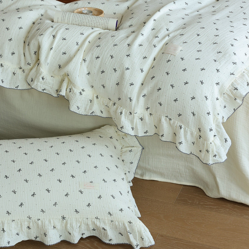 Detail image showing the ruffled pillowcase and muslin duvet cover with small bow prints. Focuses on the soft, cream-colored fabric.
